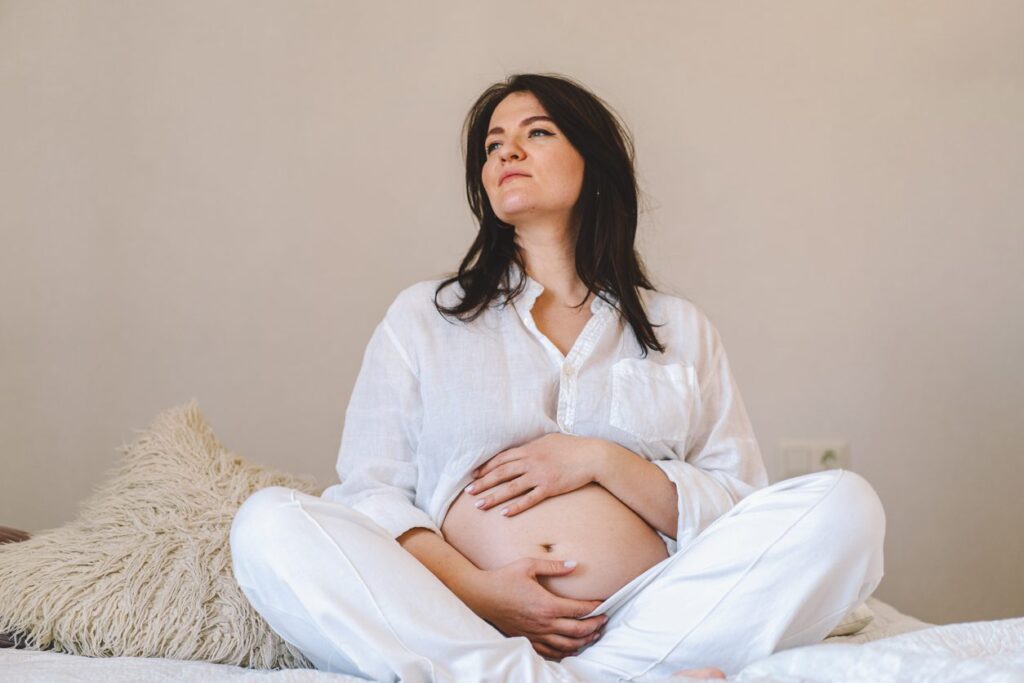 A pregnant woman sits comfortably on a bed, wearing relaxed white clothing. She gently holds her belly, embodying tranquility and connection with the child